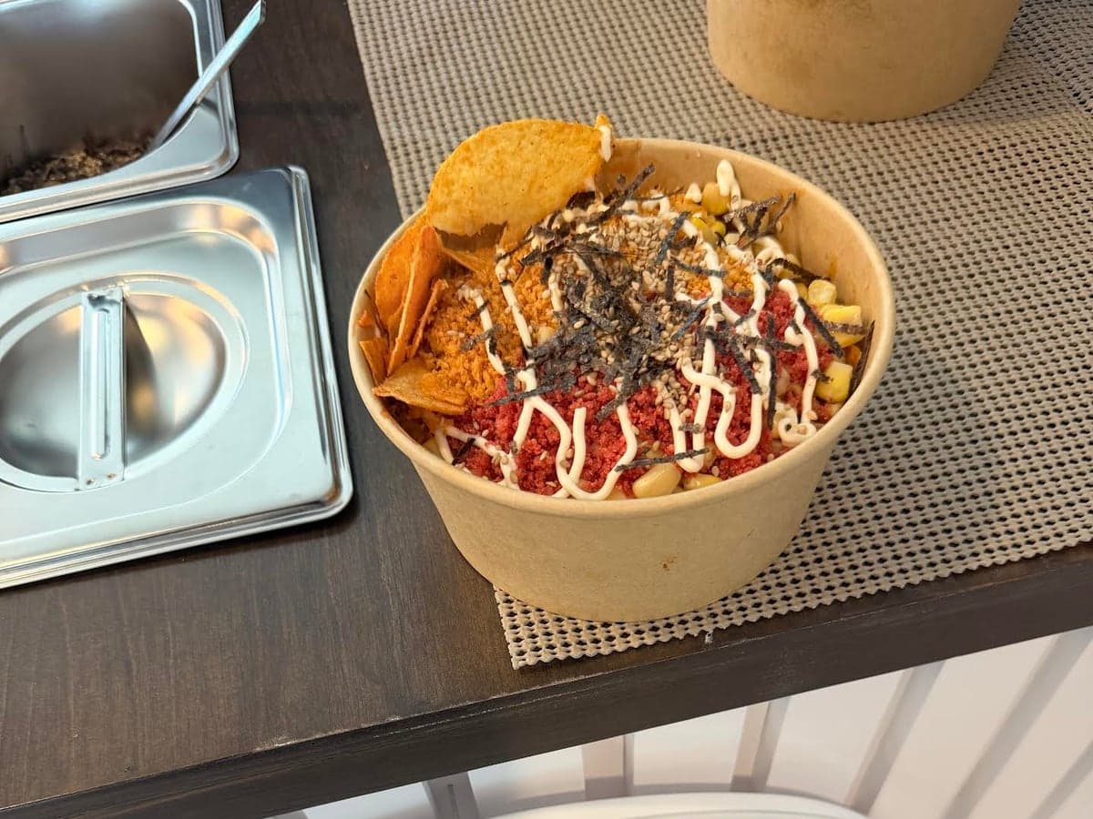 A kraft bowl of saucy ramen topped with Takis dust, furikake, nori, sweetcorn and a creamy mayo drizzle, with a bag of chips at the side